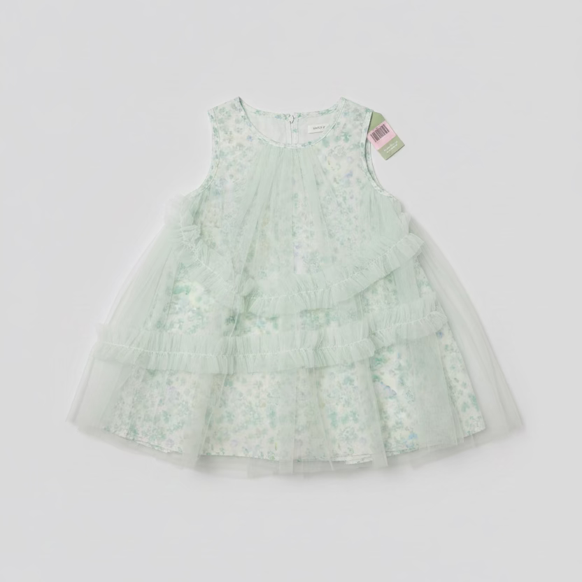 A light green and white floral tiered tulle dress laid flat on a dark wood floor, with a person's legs and shoes visible on the left side of the frame.