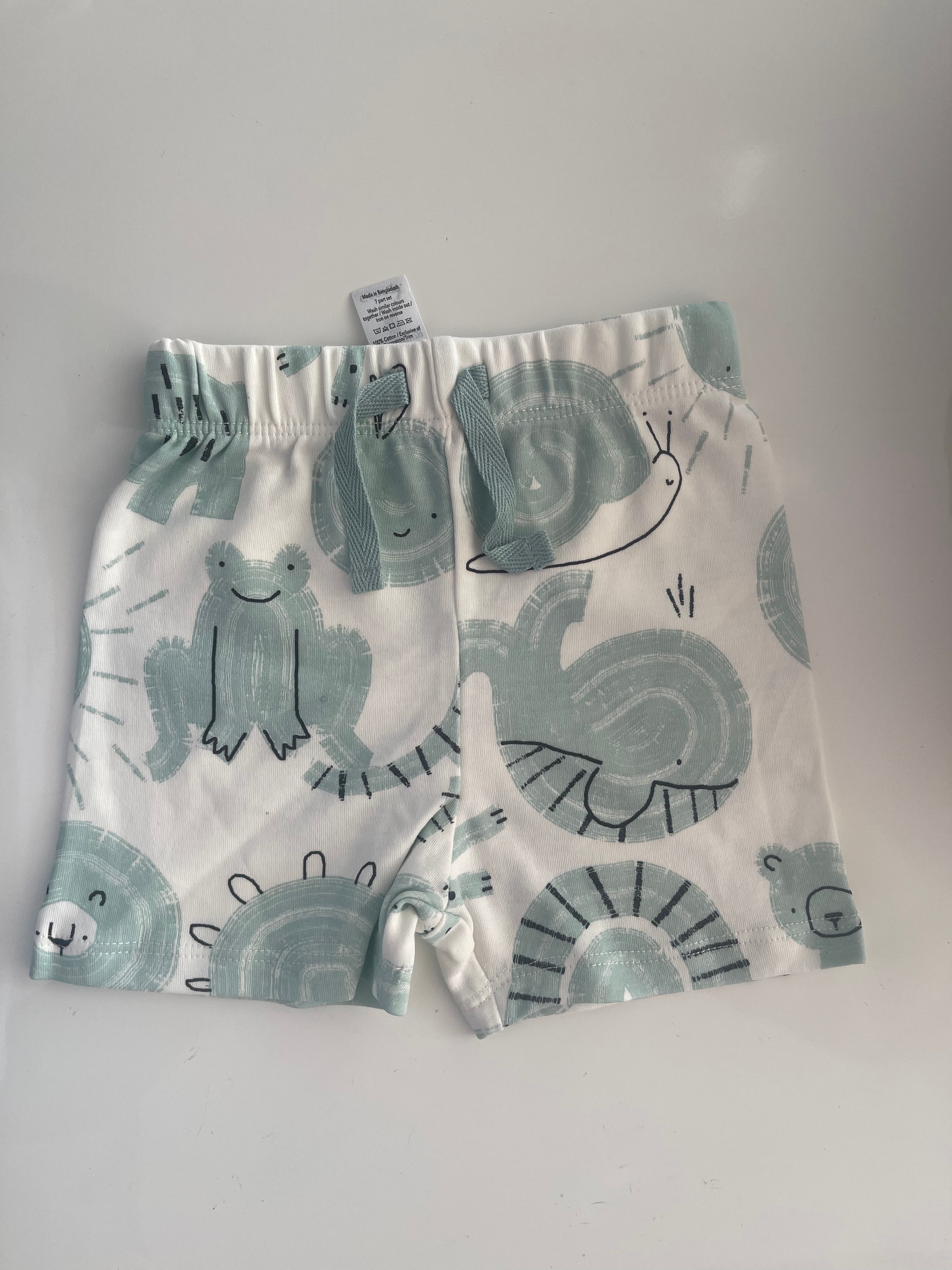 White baby shorts with a light blue-green and black animal print, featuring a frog, turtle, and sun design, laid flat on a white surface.